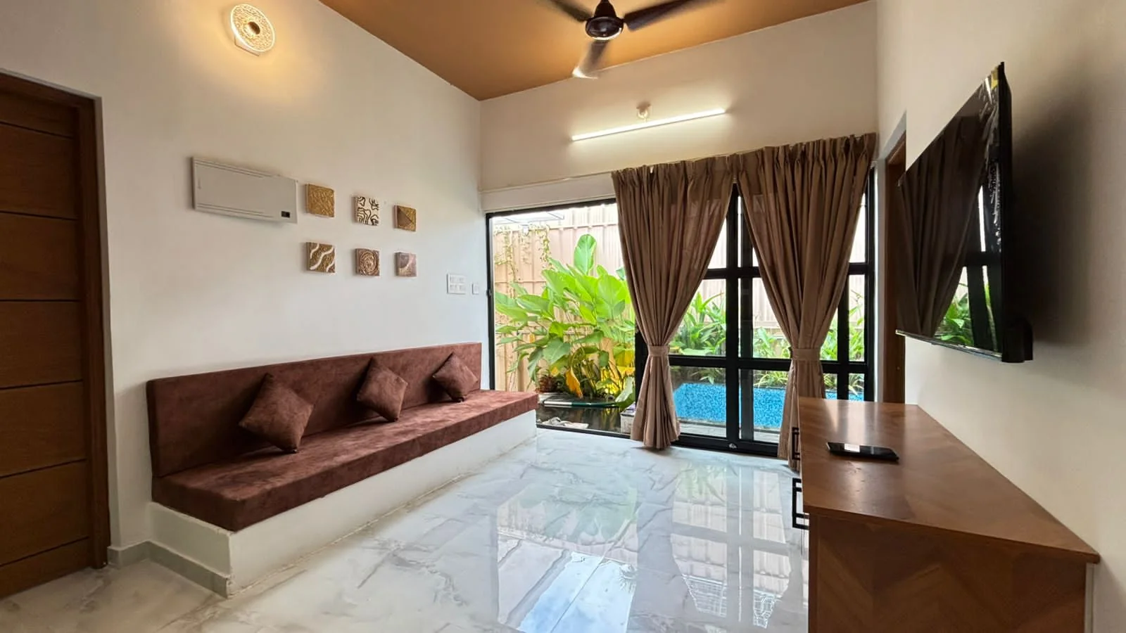 Living room with a built-in brown sofa and a glass wall opening onto tropical plants and the blue pool